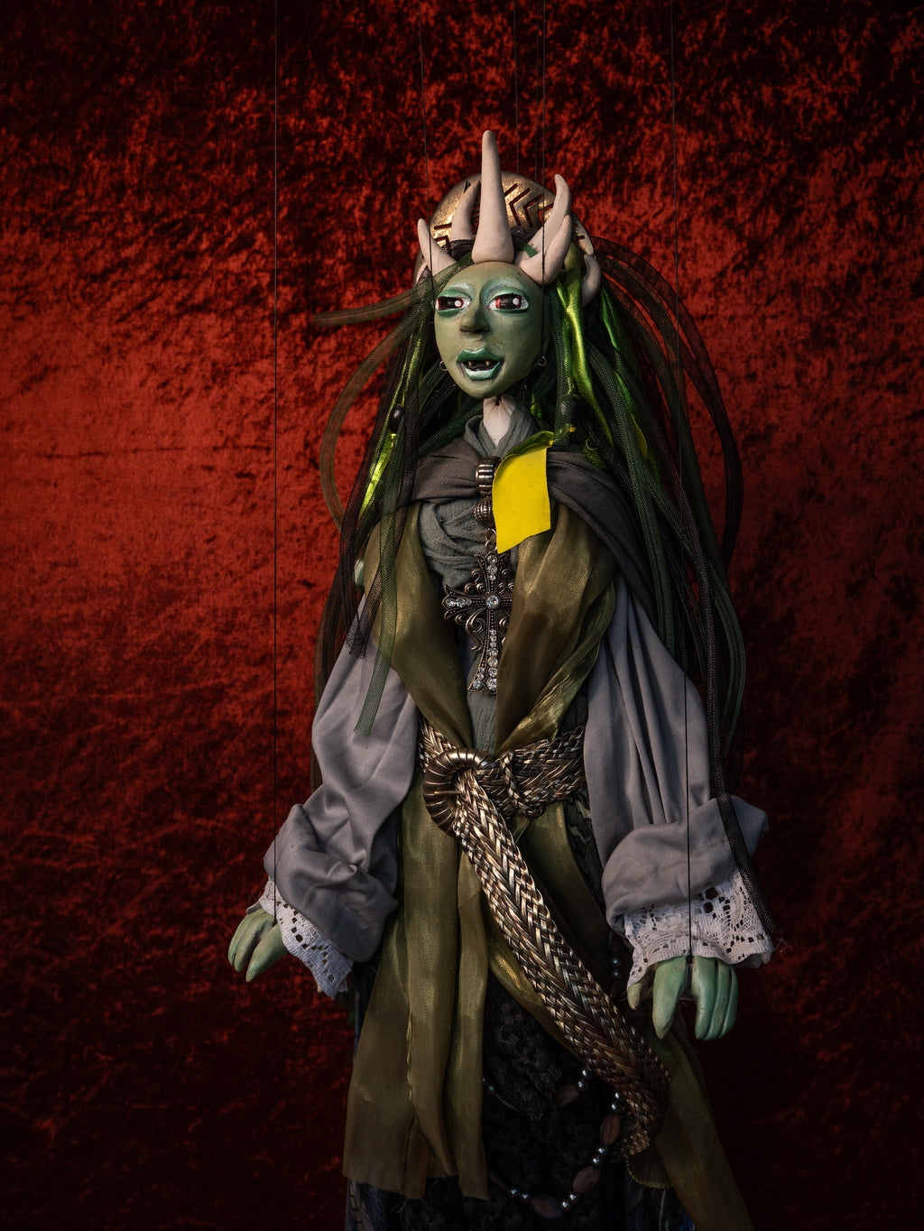 A green-skinned fantasy doll or puppet with long dark hair, horns, a gray tunic, brown outer garment, and a bright yellow chest accent, posed against a textured dark red background.