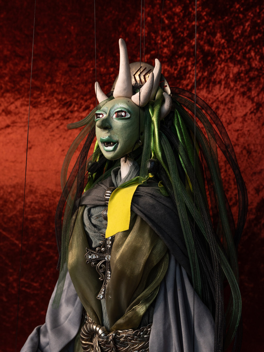 A marionette-style puppet character with a green face, long dark hair, horned headpiece, and ornate costume against a textured red background.