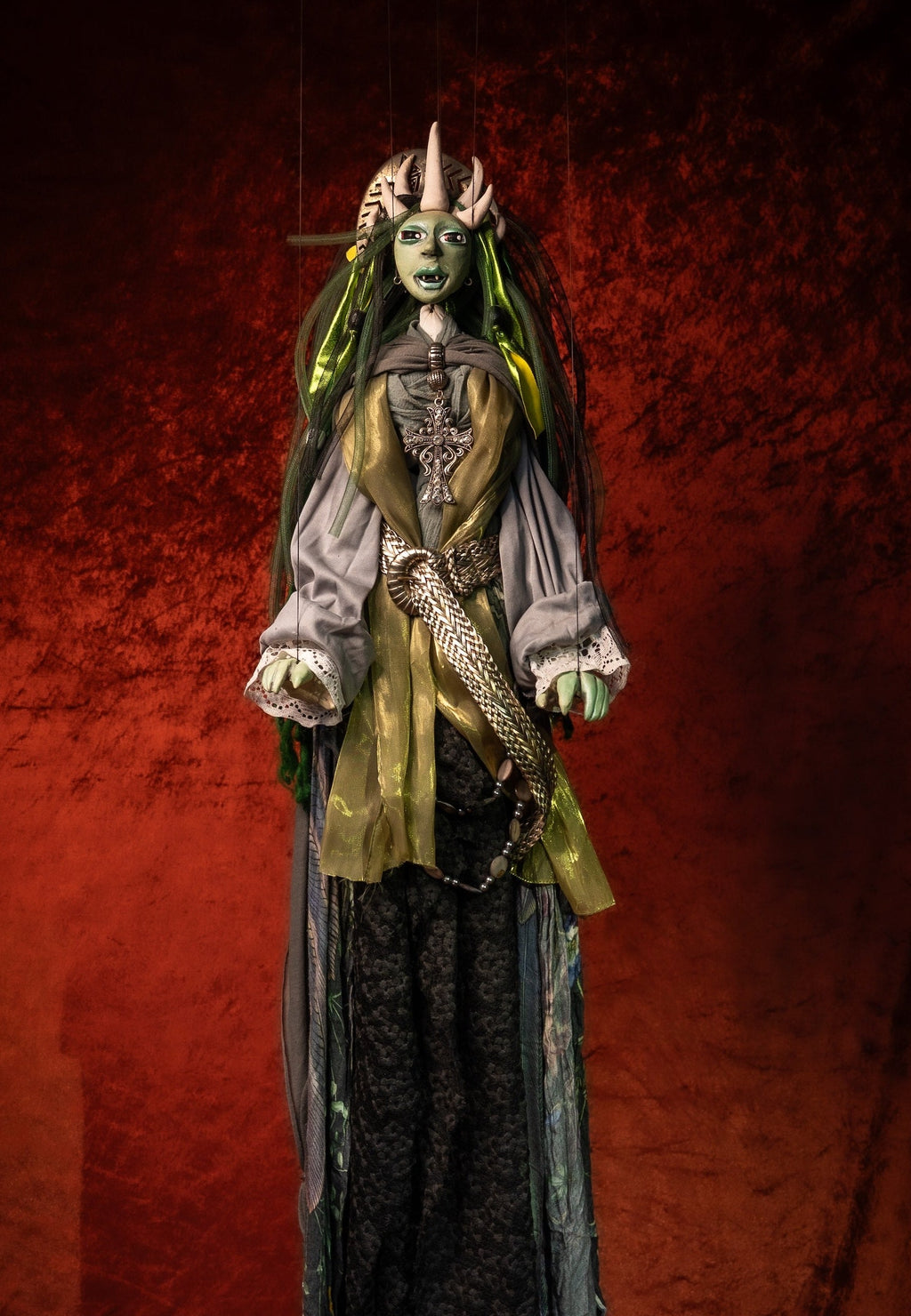 A tall, slender gothic figure or doll-like character wearing an ornate costume, standing against a dark red textured backdrop. The outfit includes a pale gray top with green accents, a metallic gold chest panel, a long dark skirt, and a decorative headdress.