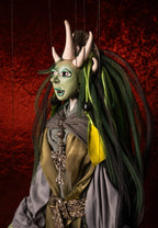 A stylized puppet or marionette figure with green skin, large pale horns, long dark green and black hair, and an ornate costume in gray, gold, and yellow, posed against a textured red background.