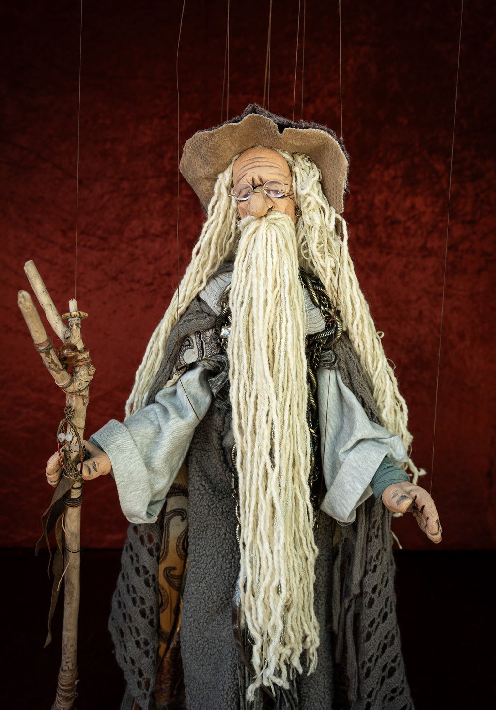 A marionette-style wizard doll with a long white beard, brown hat, gray robe, and wooden staff, suspended against a dark red textured backdrop.