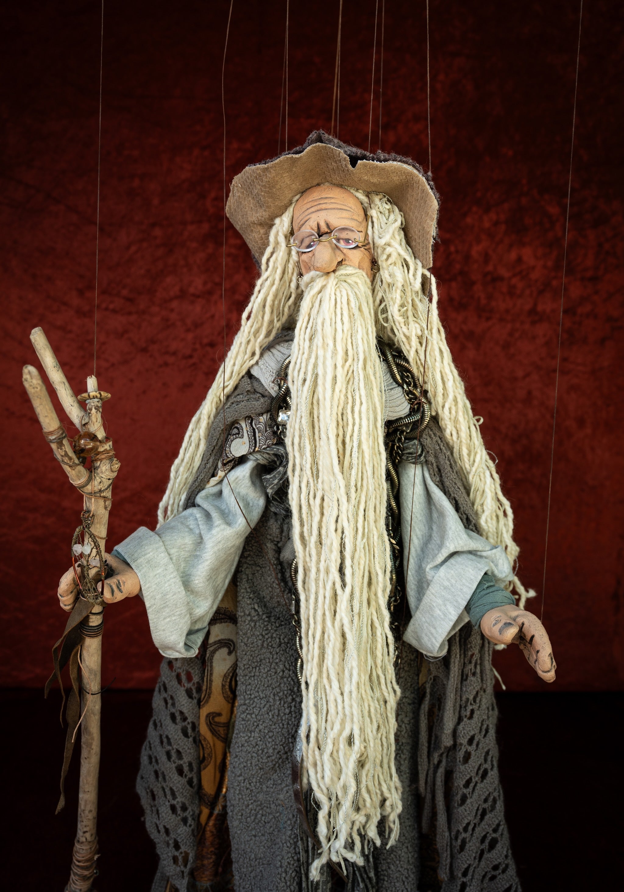 A marionette-style wizard doll with a long white beard, brown hat, gray robe, and wooden staff, suspended against a dark red textured backdrop.