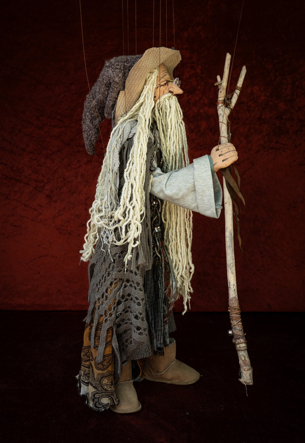 A handmade hanging puppet doll of an old wizard or storyteller, with long white yarn hair, a straw hat, tattered gray and brown garments, and a wooden staff, set against a dark red textured backdrop.