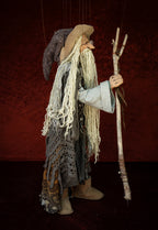 A handmade hanging puppet doll of an old wizard or storyteller, with long white yarn hair, a straw hat, tattered gray and brown garments, and a wooden staff, set against a dark red textured backdrop.