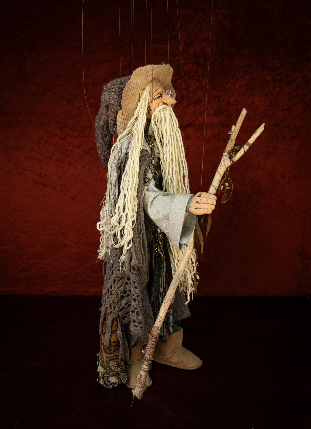 A vintage-style wooden puppet or marionette figurine of an elderly wizard or sorcerer, featuring long white yarn hair, a tan hat, and a staff, set against a dark textured red background.