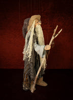 A vintage-style wooden puppet or marionette figurine of an elderly wizard or sorcerer, featuring long white yarn hair, a tan hat, and a staff, set against a dark textured red background.