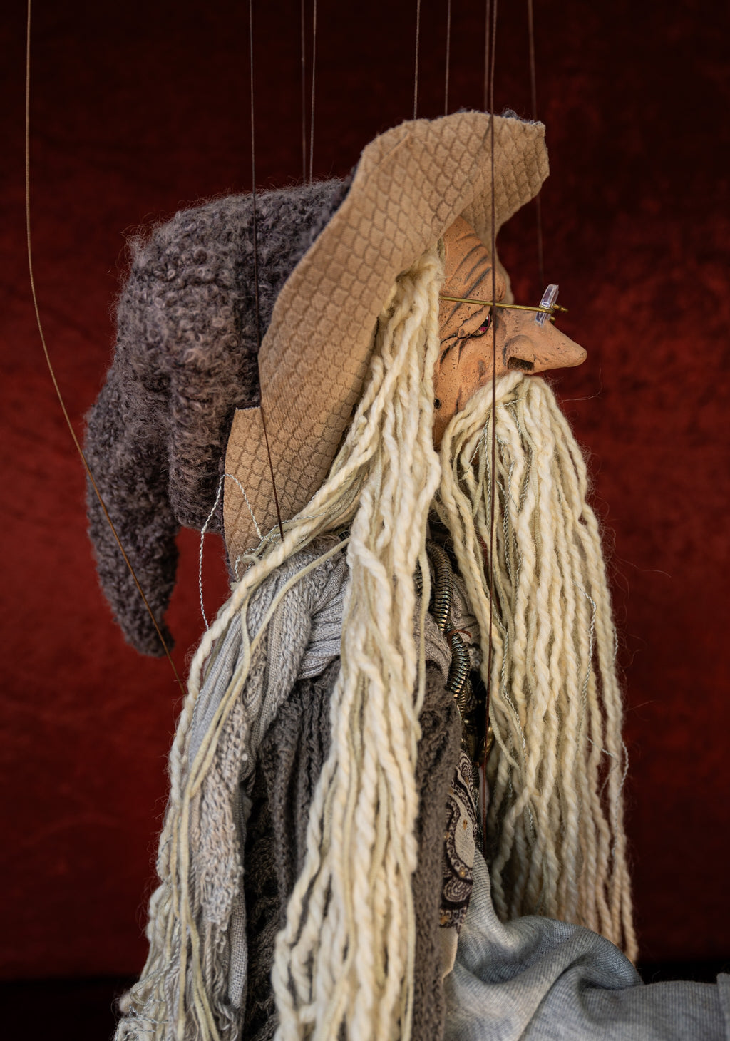 A handmade wool and yarn puppet character with long cream dreadlocks, a tan hat, and a dark gray hat, shown against a dark red textured background.