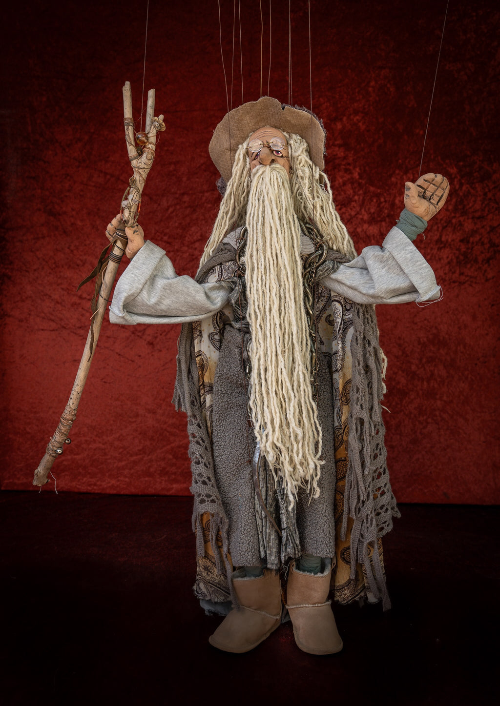 A handmade marionette doll dressed as an elderly wizard or storyteller, holding a rustic staff, set against a dark red textured backdrop.