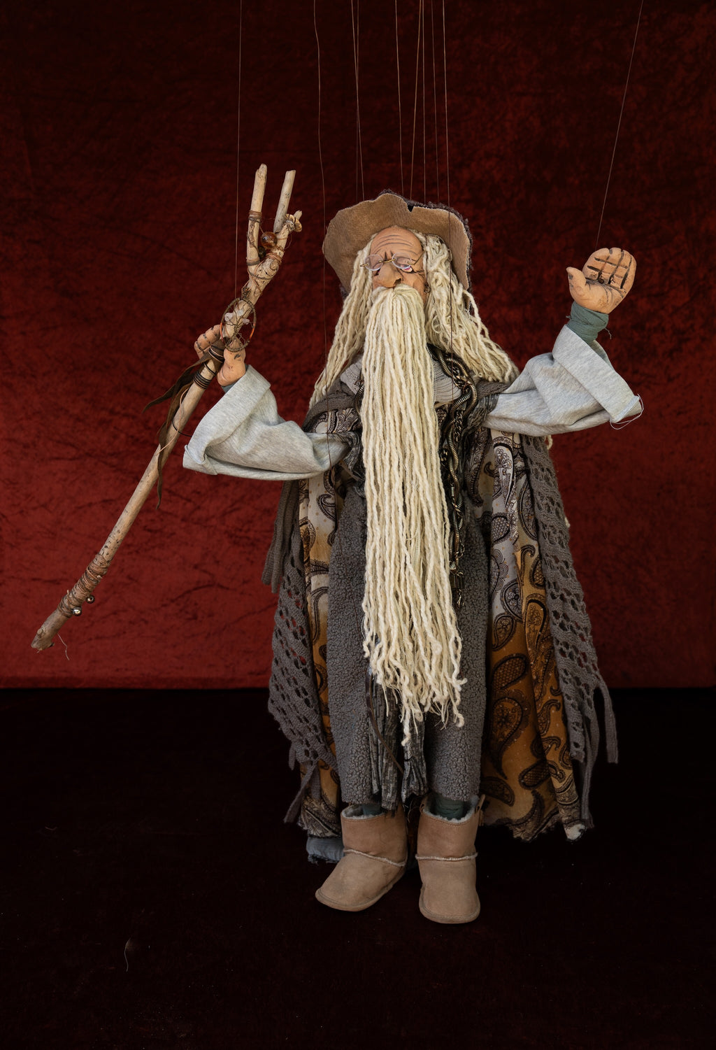 A handmade marionette-style wizard doll with a long white beard, wearing a brown hat, gray robe, and patterned cloak, holding a staff against a dark red textured backdrop.
