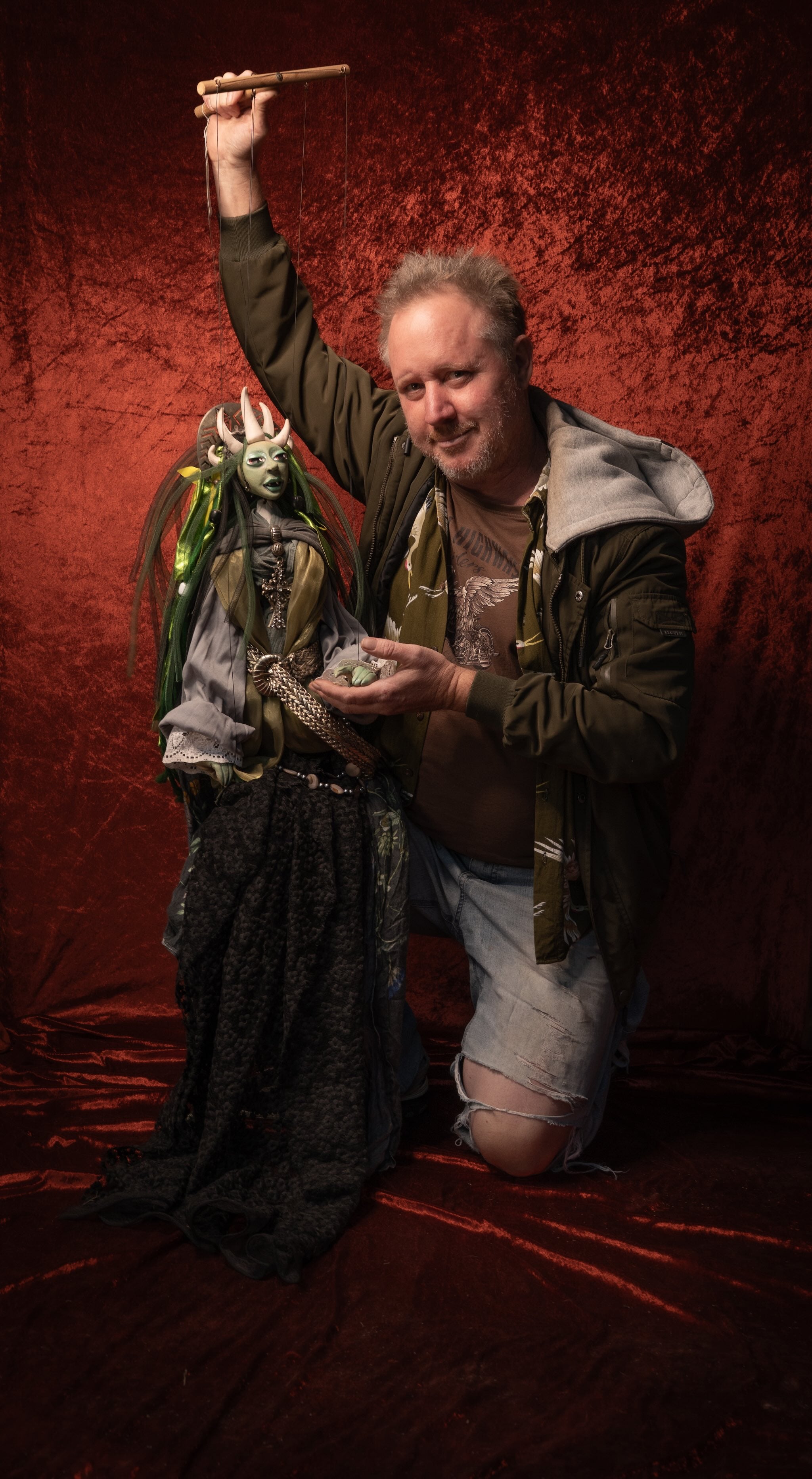 Sol Puppetman kneels on a red textured backdrop while holding a detailed fantasy puppet with a long black dress and green headpiece. He holds the crosspiece in one hand and supports the doll with the other.
